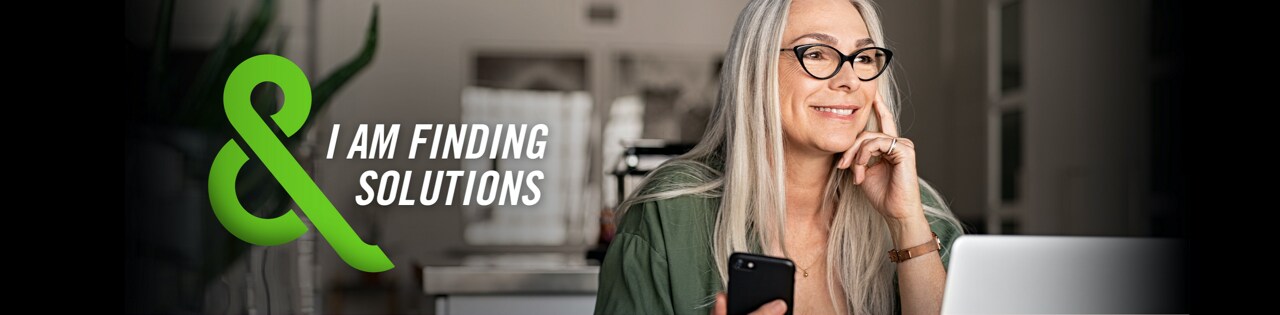 Image of a woman sitting at a laptop and holding a mobile phone with text that says – I AM FINDING SOLUTIONS